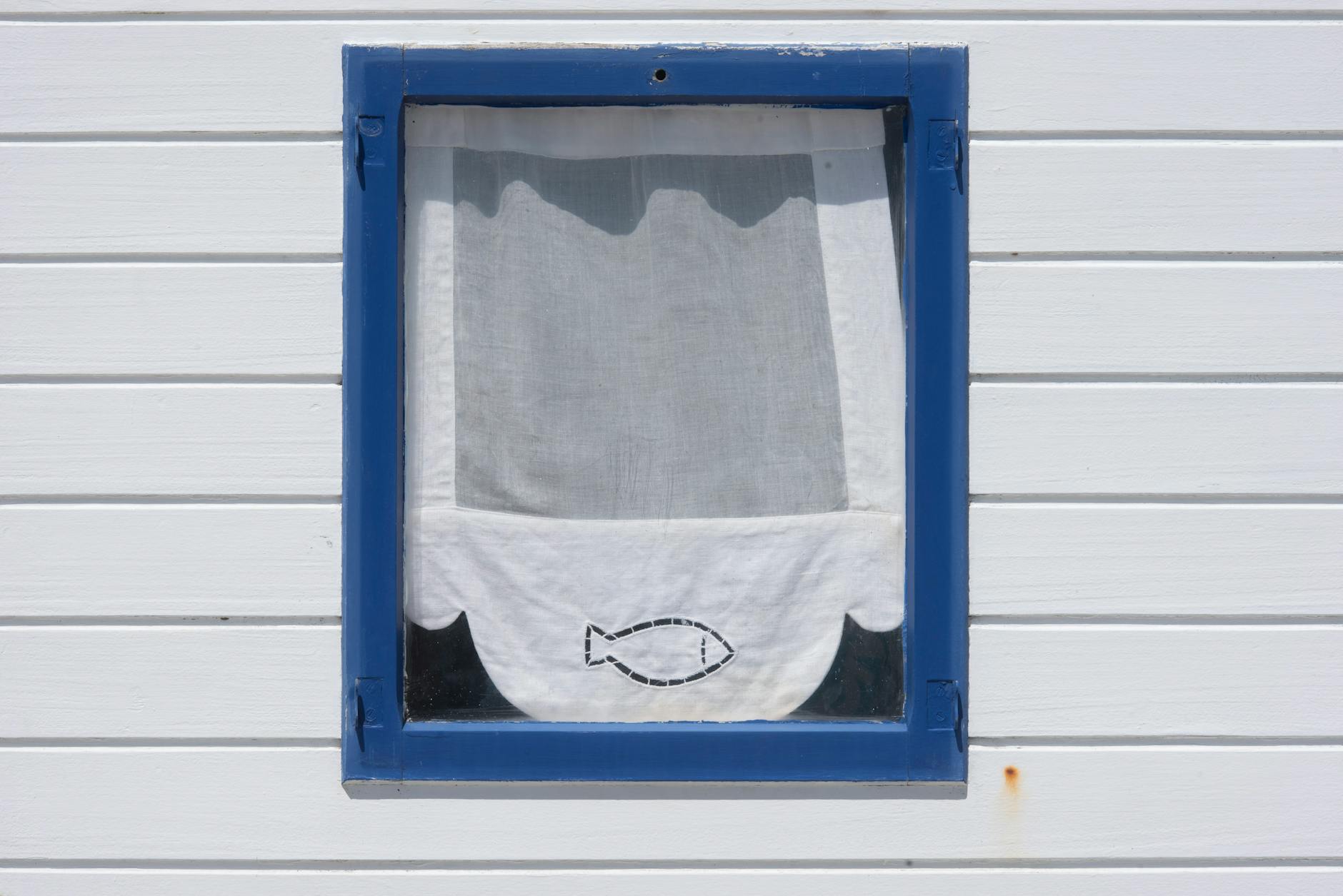 Niebieskie okno z zasłoną w ryby na białej, drewnianej elewacji